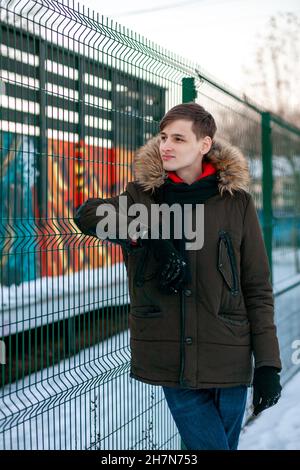 Ein junger Mann in einer Winterjacke mit Fell steht neben dem Gitterzaun und schaut dahinter. Stockfoto