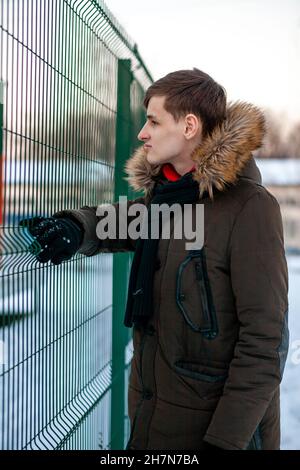 Ein junger Mann in einer Winterjacke mit Fell steht neben dem Gitterzaun und schaut dahinter. Stockfoto