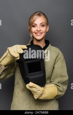 Happy blonde Schweißer in Handschuhen halten Helm und Blick auf Kamera isoliert auf grau Stockfoto