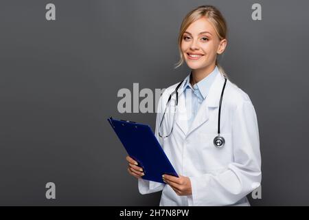 Glücklicher junger Arzt hält Zwischenablage und schaut auf Kamera isoliert auf grau Stockfoto