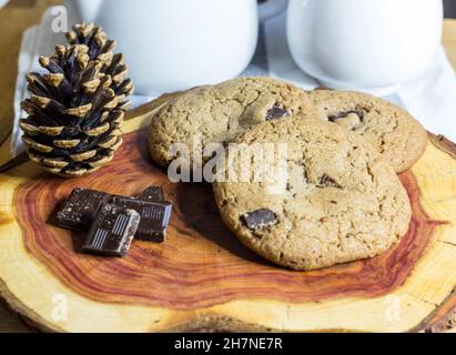 Chocolate Chip Cookies auf Holzbrett mit Schokoladenstücken und Tee mit Pinienkegel verziert serviert. Selektiver Fokus Stockfoto