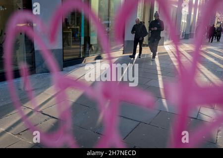 Durch die rosa Farbe der Graffiti auf dem Fenster einer öffentlichen Telefonbox gesehen, gehen Fußgänger am 23rd. November 2021 in London, England, durch das reflektierte Herbstlicht auf Moorgate in der City of London, dem Finanzdistrikt der Hauptstadt. Stockfoto