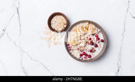 Blick von oben auf Haferflocken mit frischen Preiselbeeren, Haselnüssen, Mandeln und Bananenscheiben für ein gesundes Frühstück auf weißem Marmorboden Stockfoto