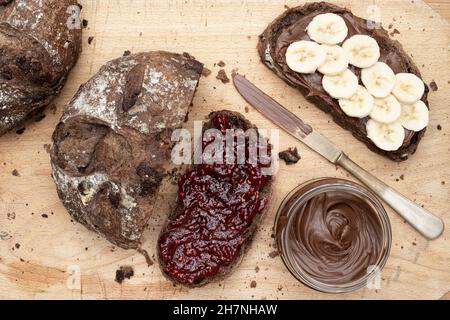 Chocolate Sourdough Bread mit Schokolade-Haselnuss-Aufstrich, Bananenscheiben und Himbeermarmelade auf einem Holzbrett Stockfoto