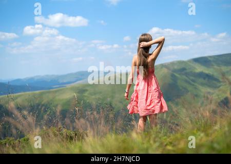Junge glückliche Frau in rotem Kleid zu Fuß auf Grasfeld an einem windigen Tag in den Bergen Sommer. Stockfoto