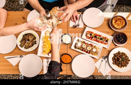 Set von Tapas, Salaten und Eintöpfen typisch für spanische Küche auf einem Holztisch, bei dem die Hände einiger Menschen in einer Restaurantbar in Spanien toasten. Stockfoto