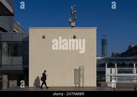 Ein japanischer Büroangestellter oder Salaryman überprüft sein Telefon, während er an einem Gebäude mit CCTV-Kameras auf dem Dach vorbei geht. Shinjuku, Tokio, Japan. Stockfoto