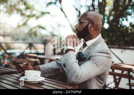 Ein modischer, reifer, kahlköpfiger afroamerikanischer Geschäftsmann mit einem schwarzen, gepflegten Bart, in einem eleganten karierten Kostüm, mit einem Smartphone in der Hand Stockfoto