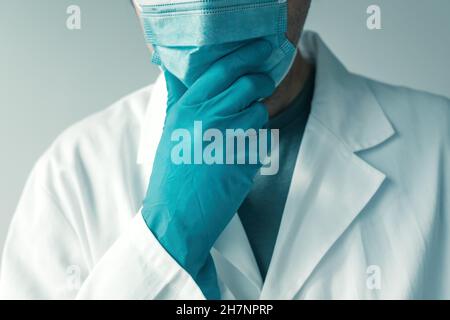 Arzt mit Schutzhandschuhen und Gesichtsmaske, die Hand am Kinn hält und denkt, selektiver Fokus Stockfoto