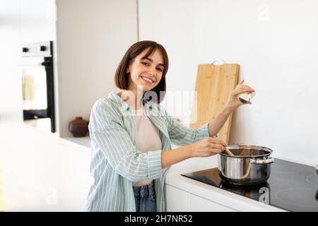 Glückliche junge Frau, die der Suppe Salz zugab und lächelte, stand am Herd in einer hellen modernen Küche Stockfoto