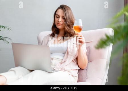 Junge, schöne Frau, die auf einem Sessel sitzt und Roséwein trinkt und einen Laptop benutzt. Kaukasische entspannte Frau, die sich einen Film auf dem Laptop ansieht oder online macht Stockfoto