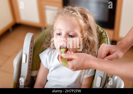 Schöne kleine blonde Mädchen essen aus Löffel isst Brei. Weibliche Mutter die Hände füttern Tochter Baby Kind kaukasischen Mädchen in High Chair AT Stockfoto