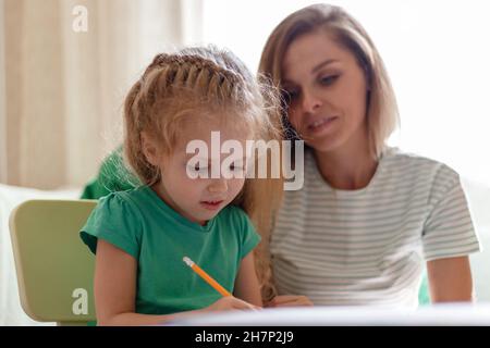 Mutter hilft Vorschulmädchen Tochter Hausaufgaben zu machen. Kaukasische blonde intelligente Kind Kind 5 Jahre Hausaufgaben machen Lernen, mit Mama 30s zusammen zu schreiben Stockfoto