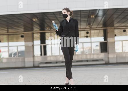 Schöne Geschäftsfrau in einem schwarzen Anzug in einer schwarzen medizinischen Maske und Handschuhe in der Stadt in Quarantäne und Isolation. Pandemie COVID-19. Bild mit sele Stockfoto