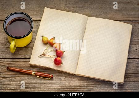 Altes leeres Notizbuch oder Reisejournal mit einem Becher aus Metall mit Tee, Stift und Krabbenäpfeln gegen rustikales Holz Stockfoto