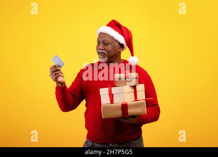 Fröhlicher älterer schwarzer Mann im Weihnachtsmütze, der Weihnachtsgeschenke und Kreditkarte hält und Weihnachtseinkäufe auf orangefarbenem Hintergrund macht Stockfoto