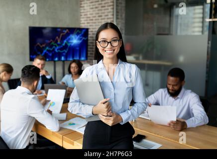 Lächelnde Asiatische Mitarbeiterin, Die Während Eines Treffens Mit Kollegen Im Innenbüro Posiert Stockfoto