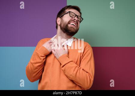 Der kaukasische bärtige Mann leidet an Allergien, er kratzt ärgerlich am Hals, schließt die Augen und entblößt die Zähne. Farbenfroher Hintergrund. Stockfoto