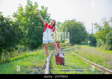 Fröhliche Frau mit Hut geht mit einem Koffer auf den Schienen Stockfoto