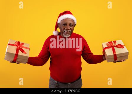 Glücklicher älterer schwarzer Mann in rotem Pullover und Weihnachtsmütze mit Weihnachtsgeschenkschachteln in beiden Händen, der große Weihnachtsverkäufe anbietet Stockfoto