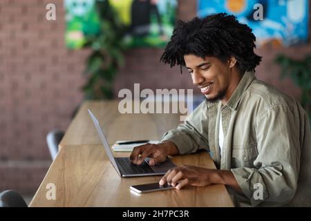 Junger afroamerikanischer Geschäftsmann, der im Café mit dem Smartphone arbeitete, während er mit dem Laptop arbeitete Stockfoto