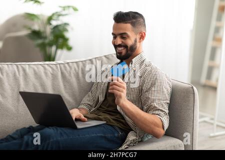 Hübscher junger arabischer Mann mit Laptop und Kreditkarte sitzt auf dem Sofa und kauft zu Hause im Webshop ein Stockfoto