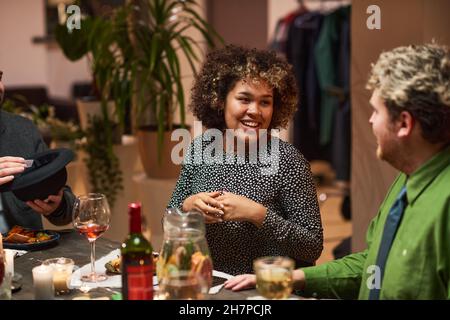 Glückliche Afrikanerin, die beim festlichen Abendessen lachend und mit ihren Freunden am Esstisch sprach Stockfoto