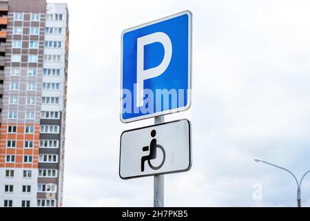 Blau-weißes Parkschild mit Rollstuhl für Menschen mit Behinderungen vor Himmel und Stadtgebäuden Hintergrund. Stockfoto
