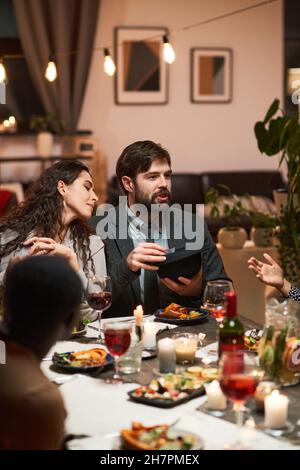 Junge Leute sitzen am festlichen Esstisch und unterhalten sich, sie essen im Wohnzimmer Stockfoto