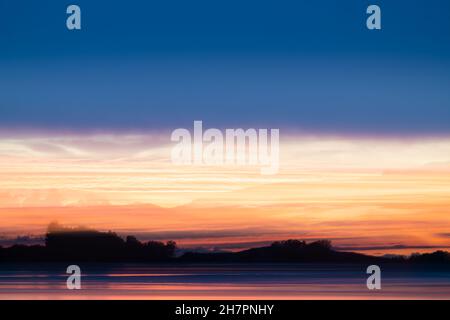 Abstraktes Küstenmotiv mit Langzeitbelichtung für intensiven Farbhintergrund mit orangefarbenem Farbton, der einen Hauch von Landsilhouette und Wolken am Horizont zeigt. Stockfoto