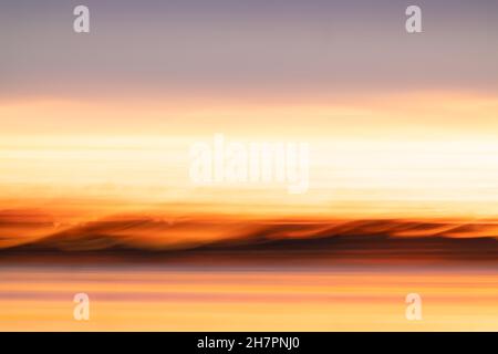 Abstraktes Küstenmotiv mit Langzeitbelichtung für intensiven Farbhintergrund mit orangefarbenem Farbton, der einen Hauch von Landsilhouette und Wolken am Horizont zeigt. Stockfoto