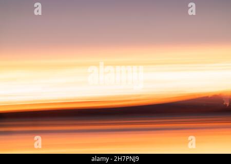 Abstraktes Küstenmotiv mit Langzeitbelichtung für intensiven Farbhintergrund mit orangefarbenem Farbton, der einen Hauch von Landsilhouette und Wolken am Horizont zeigt. Stockfoto