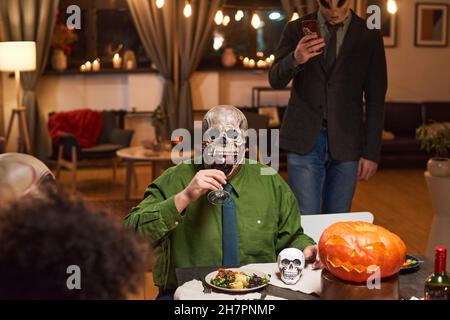 Porträt eines Mannes in einer unheimlichen Maske, der Rotwein trinkt, während er am Tisch sitzt und mit seinen Freunden eine Halloween-Party feiert Stockfoto