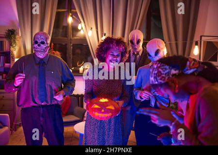 Afrikanische Frau hält beleuchteten Kürbis und Spaß zusammen mit ihren Freunden auf Halloween-Party zu Hause Stockfoto
