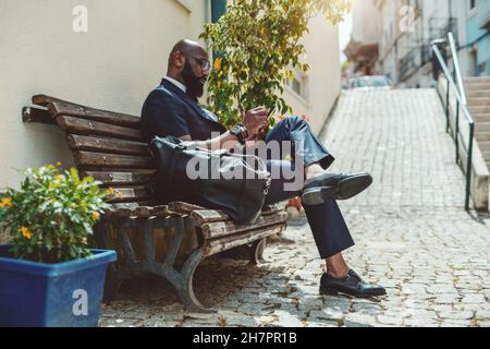 Ein hübscher, reifer bärtiger, kahlköpfiger Schwarzmann-Unternehmer in einem eleganten Outfit sitzt auf einer hölzernen Straßenbank auf dem Bürgersteig und benutzt sein Smartphone; A Stockfoto