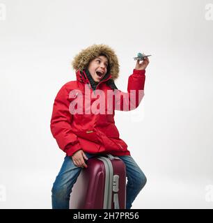 Entzückender Schuljunge in warmer roter Daunenjacke mit Kapuze, sitzt auf dem Gepäck und spielt mit einem Spielzeug-Modell eines Flugzeugs, imitiert den Flug am Himmel Stockfoto