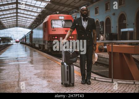 Ein modischer, reifer, kahlköpfiger afroamerikanischer Unternehmer in einem eleganten Anzug mit Weste und Fliege steht auf einer Plattform eines Bahnhofs d Stockfoto