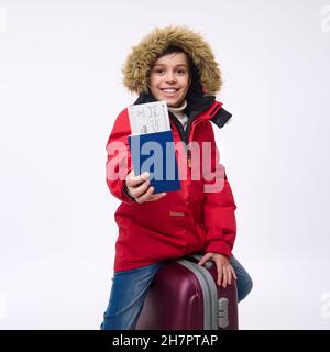 Fröhlicher Jugendlicher, Junge im Schulalter in hellrotem Parka, der auf seinem Gepäck sitzt, lächelt toothy lächelt und schaut auf die Kamera und zeigt Pass mit dem Einsteigen Stockfoto