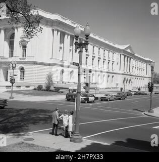 1960s, historisch, ein junger Mann und zwei junge Damen warten darauf, die Constitution Avenue in Washington DC, USA, der Hauptstadt des Landes zu überqueren. An der gegenüberliegenden Kreuzung warten Autos der Ära auf die Straße, ein gelbes Taxi und ein US-Postauto. Große US-Regierungsbehörden befinden sich in vielen der großen neoklassizistischen Säulengebäude der Straße. Ursprünglich einfach als B St. NW bekannt, als das Gebiet Ende 1930s saniert wurde, wurde vorgeschlagen, es zu Ehren des dritten US-Präsidenten Thomas Jefferson und des Gründervaters in Constitution Avenue umzubenennen, was 1943 geschah. Stockfoto