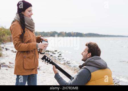 Lächelnde Frau, die aus Thermoskannen in der Nähe des Mannes, der am Flussufer akustische Gitarre spielt, trinkt Stockfoto