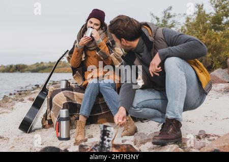 Junge Frau trinkt aus dem Thermobecher in der Nähe von Freund und Lagerfeuer an der Flussküste Stockfoto