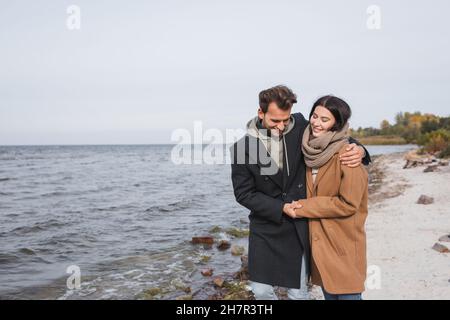 Fröhliches Paar in herbstlichen Mänteln, das sich während des Spazierens am Flussufer die Hände hält Stockfoto