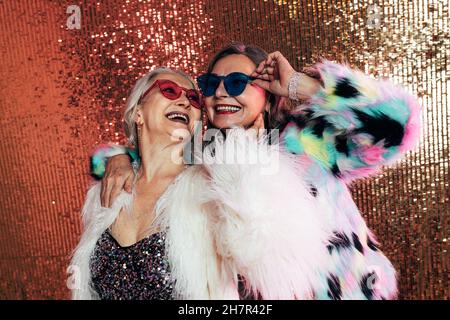 Zwei Frauen tragen Brillen und Pelzmäntel. Ältere Frauen, die vor glitzerndem Hintergrund lachen. Stockfoto