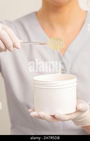 Flüssige gelbe Zuckerpaste zur Enthaarung auf einem Stock fließt in den Becher in den Händen einer Kosmetikerin Stockfoto