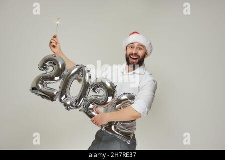 Ein lächelnder Mann in einem roten Samthut hält mit einer Hand silberne Luftballons in der Form von 2022 und mit der anderen Hand einen Funker. Ein glücklicher Kerl Stockfoto