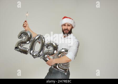 Ein Mann in einem roten Samthut hält mit einer Hand silberne Ballons in Form von 2022 und mit der anderen Hand einen Funker. Ein glücklicher Typ mit einem b Stockfoto