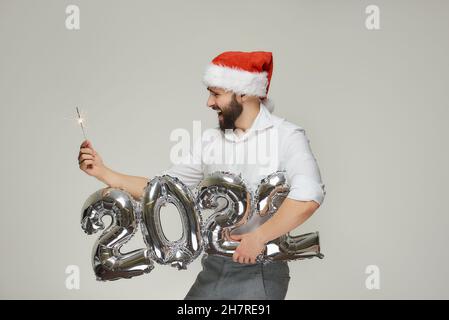 Ein Mann in einem roten Samthut hält mit einer Hand silberne Ballons in Form von 2022 und mit der anderen Hand einen Funker. Ein Kerl mit einem Bart ich Stockfoto