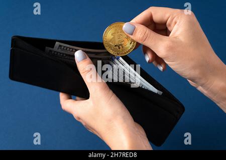Goldene Bitcoin-Münze in den Händen eines Mädchens mit einer Geldbörse mit Dollarnoten auf blauem Hintergrund. Nahaufnahme Stockfoto