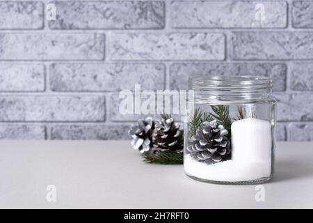 Weiße Kerze in einer Glasvase mit Tannenzapfen und Fichtenzweigen auf schwarzem und grauem Hintergrund. Das Konzept der Kerzenständer im Innenraum des KI Stockfoto