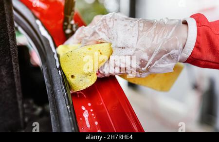 Junge Frau putzt den hinteren Teil ihres Autos, Nahaufnahme Detail auf der Hand im Handschuh mit gelben Schwamm Stockfoto